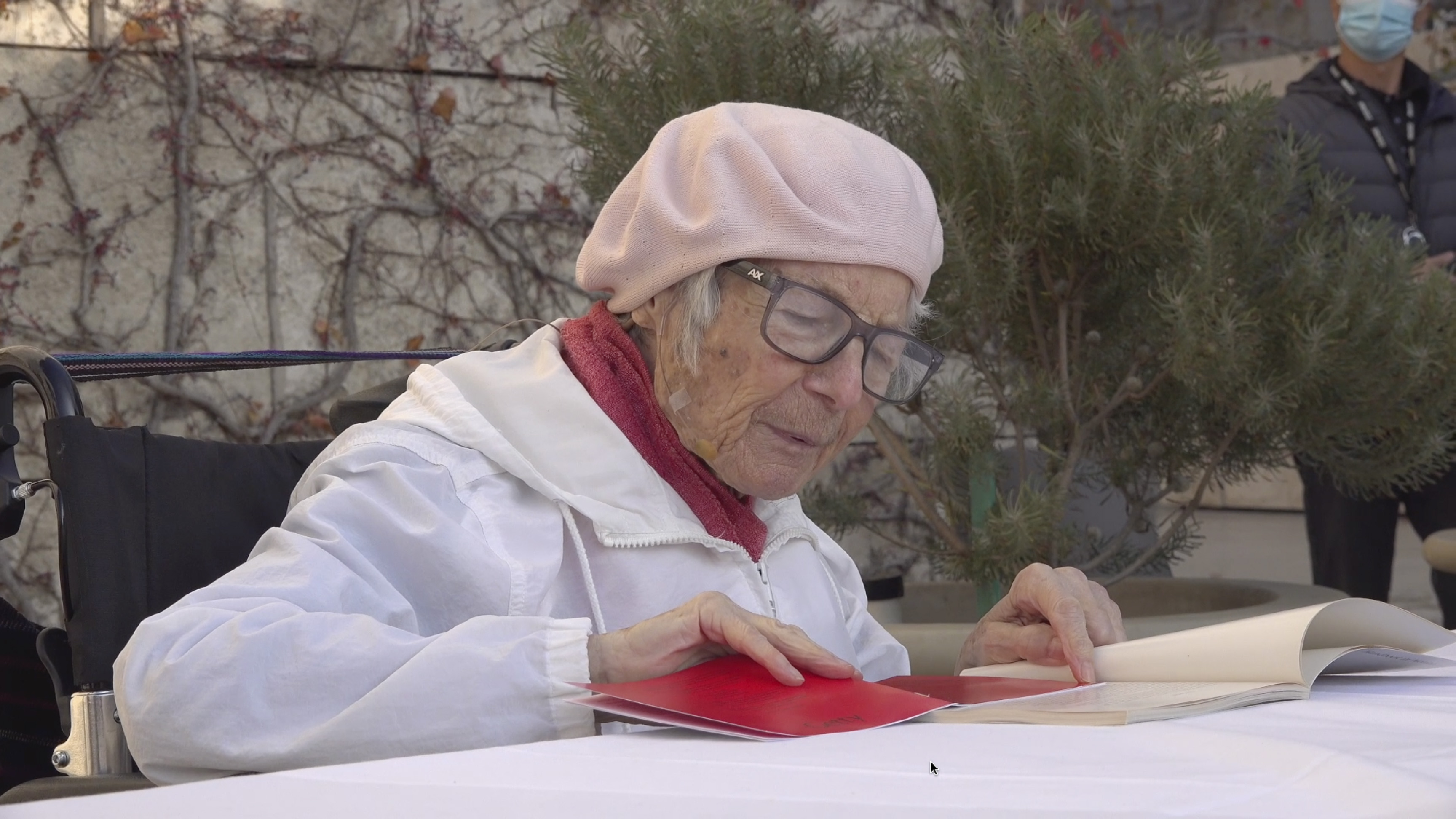 Color video showing elderly woman wearing glasses, light pink beret, white jacket, and a barely visible headset microphone sits at a table set up outdoors on museum plaza. She reads excerpts of personal narratives from a book that she wrote, and the audience shows their appreciation with occasional gentle laughter.