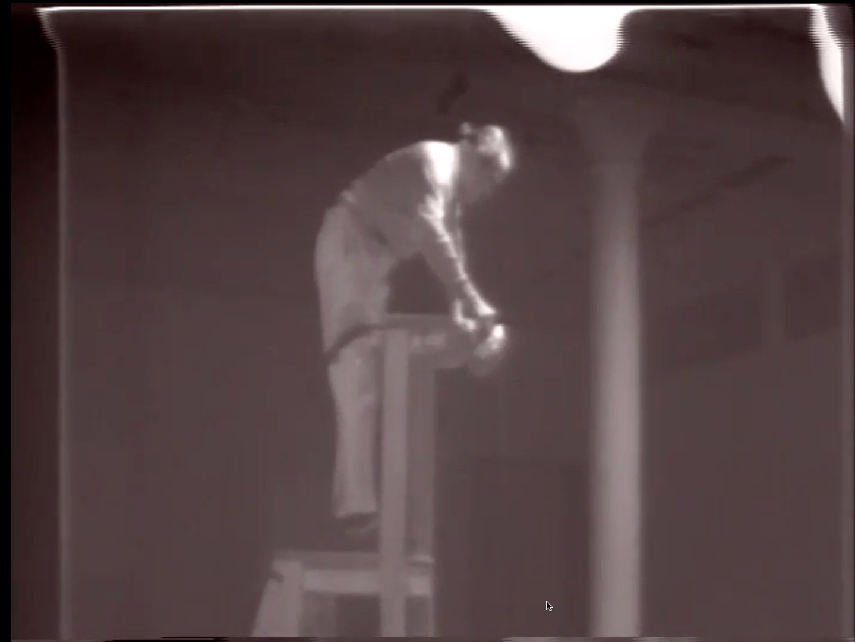 Black and white video of man placing chair, bowl, pitcher of water, and rolling wooden ladder at the center of a room in front of an audience sat on the floor just below view of camera. The man announces “Drip Music,” and then steps onto the ladder and audibly pours water from the pitcher into the bowl. The camera often zooms in to follow the movements of objects in the man’s hands as well as the stream of water.  