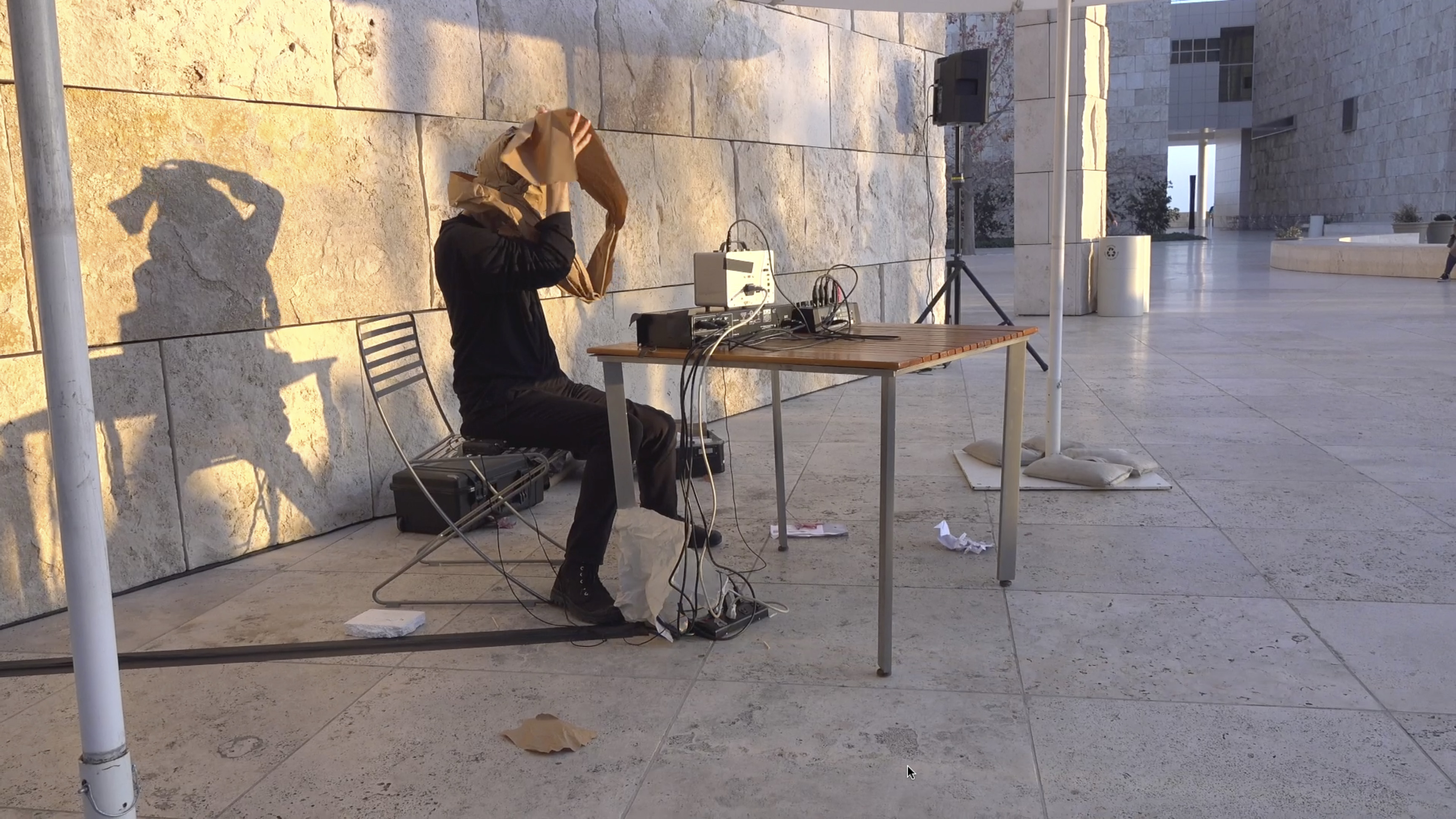 Color video showing man wearing all black sitting outdoors at a table with electronic sound equipment on it. He wraps lengths of butcher paper around his head so that eventually he cannot see, while around his neck drapes a contact microphone that amplifies the sounds in a grating manner. Once his head is completely wrapped, he sits in near silence.