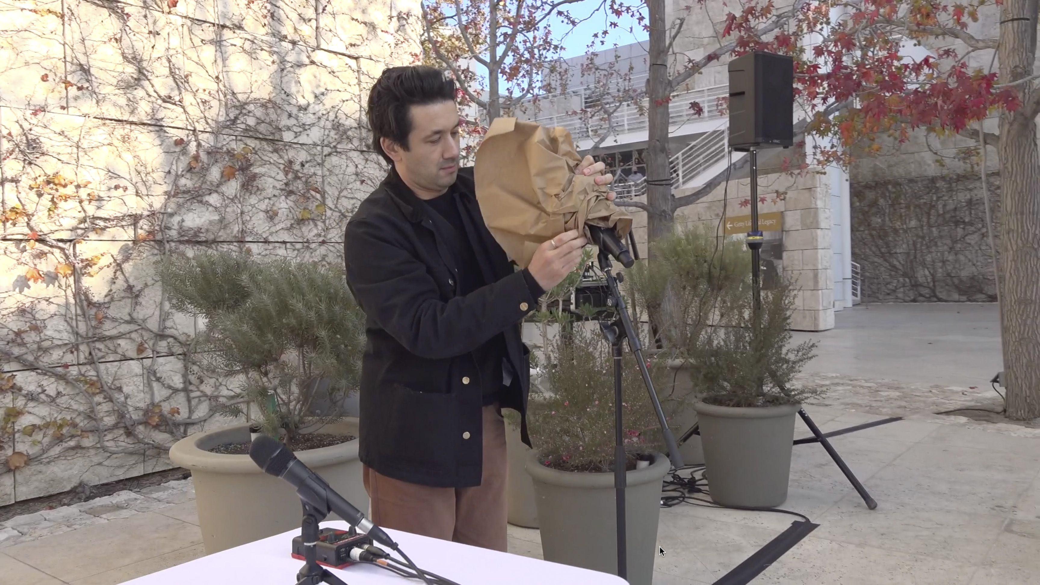 Color video showing a live microphone on stand set up outdoors on museum plaza. Man approaches with a length of butcher paper which he quickly wraps around the microphone, thereby making loud, crunching sounds. He stands aside to listen for further sounds; the breeze and a quiet feedback tone can be heard.