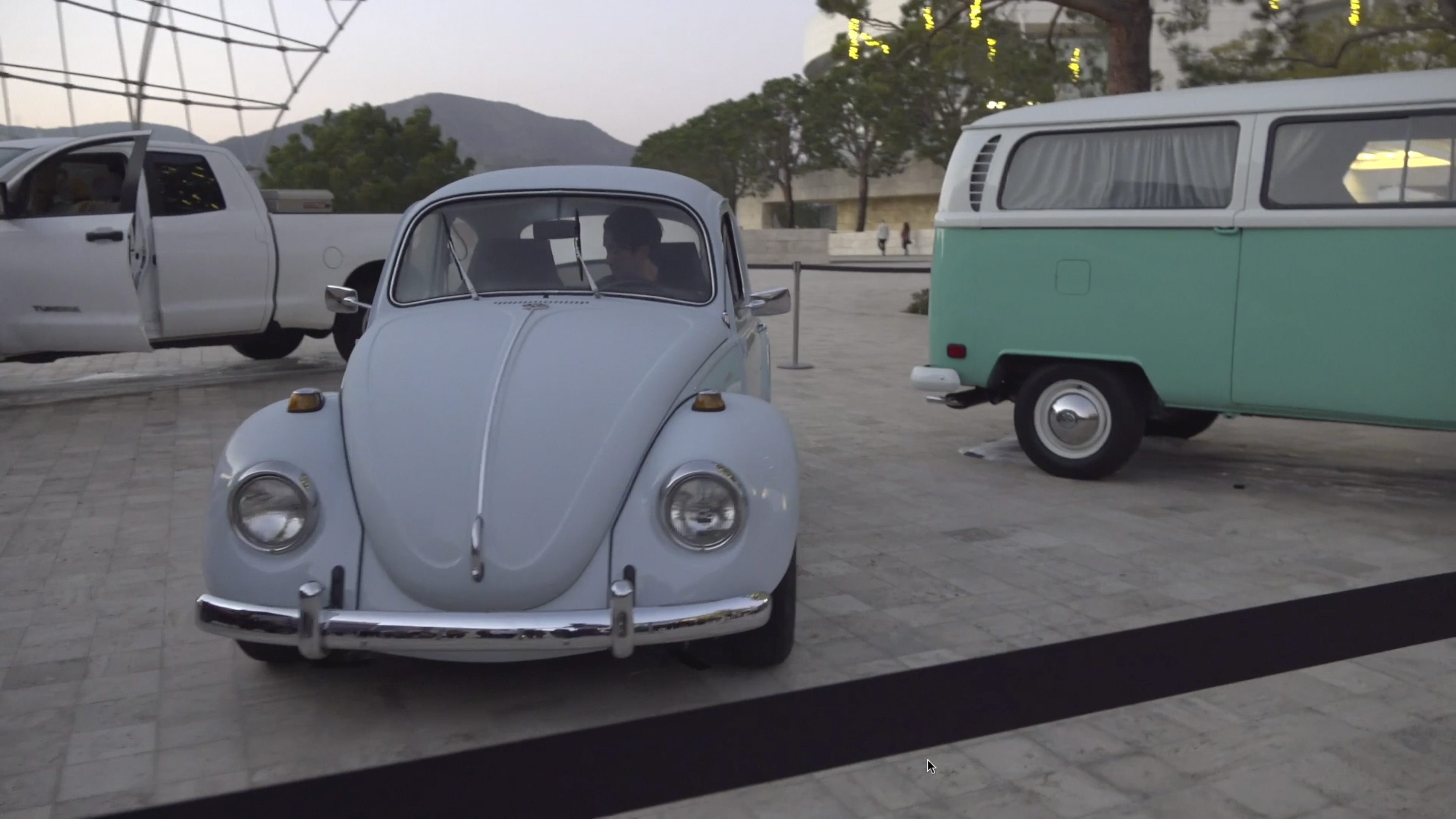 Color video showing four vehicles positioned outdoors on a museum plaza at sunset: a white pickup truck, small utility vehicle, Volkswagen Beetle, and Volkswagen van. One performer enters each vehicle and starts its engine. In random order, the vehicles’ engines rev, lights and radios turn on and off, windshield wipers turn on and off, doors open and close, and horns honk. The performer in the Beetle occasionally sounds a hand-operated air raid siren. The light around gradually darkens as the sun goes down.