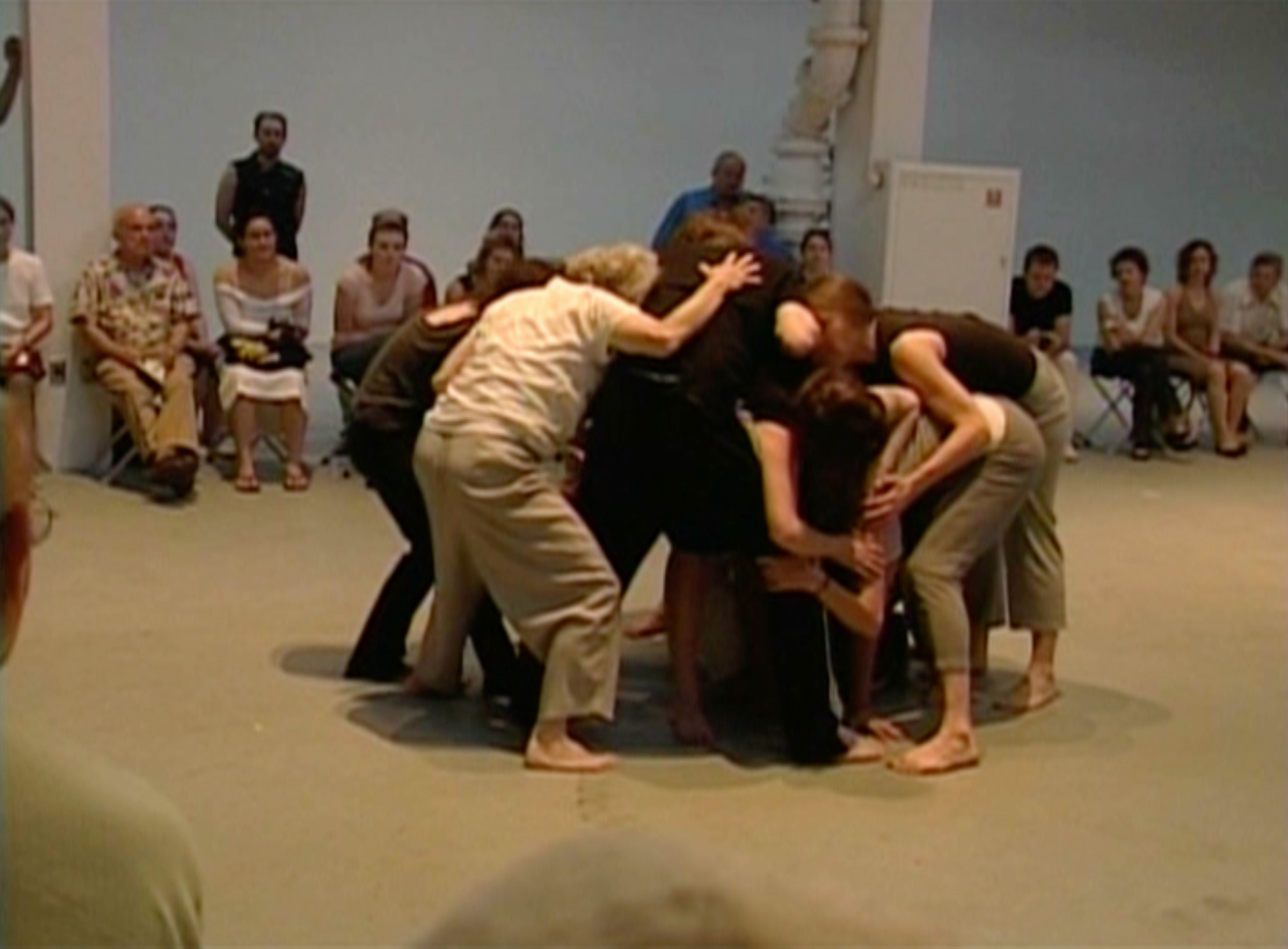 Color video recording taken inside a warehouse-like museum space. The first performance involves ten performers, including Simone Forti, grouped together in the middle of a large circle of spectators who are sitting on folding stools. The performers, who are barefoot and wearing comfortable street clothes, form a tight circle with their bodies, crouching down and holding their arms around one another’s shoulders. One performer at a time slowly and carefully climbs up and over the cluster of bodies. At one point Forti can be heard saying, “Take your time, take your time”; otherwise the event is largely silent. It concludes when the performers stand upright and break apart. The second performance, which takes place at one end of the space, involves four of the performers who now wear tennis shoes. They interact with a large, ramp-like board mounted at a 45-degree angle and with five evenly spaced, knotted ropes attached to the top of the incline. The performers hang onto the ropes as they climb up, down, and across the board, focused on their individual actions. The performance is largely silent except for the sounds of shoes and ropes making contact with the board.