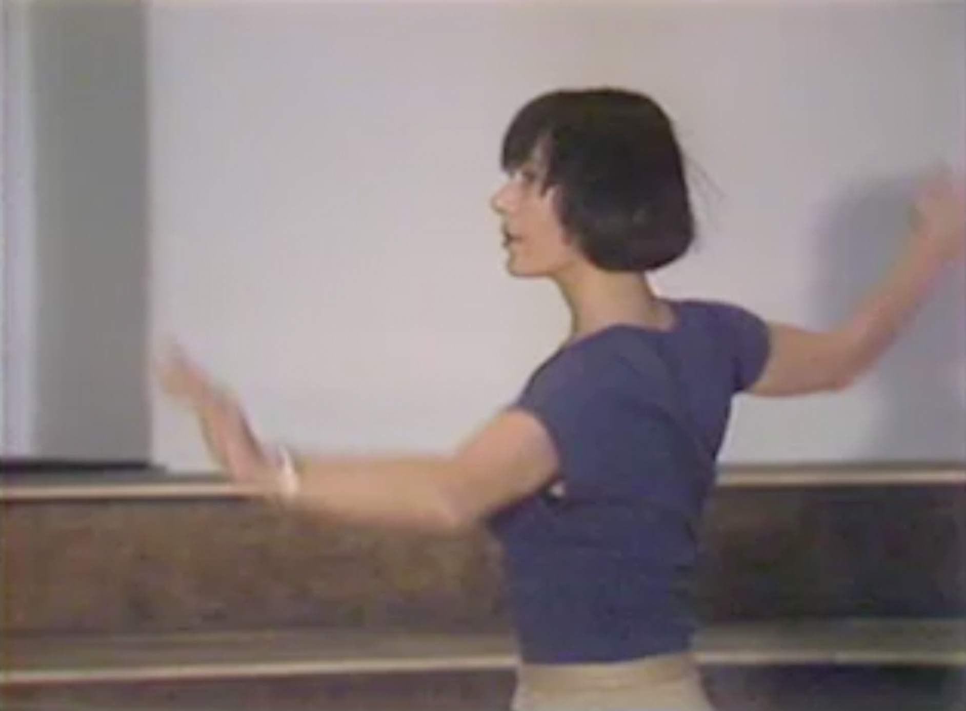 Color video featuring a male interviewer holding a microphone and a woman (Yvonne Rainer) answering questions, set in a large performance hall with dancers behind them. The video cuts to Rainer dancing alone in the hall, wearing a purple t-shirt, loose-fitting slacks, and sneakers.