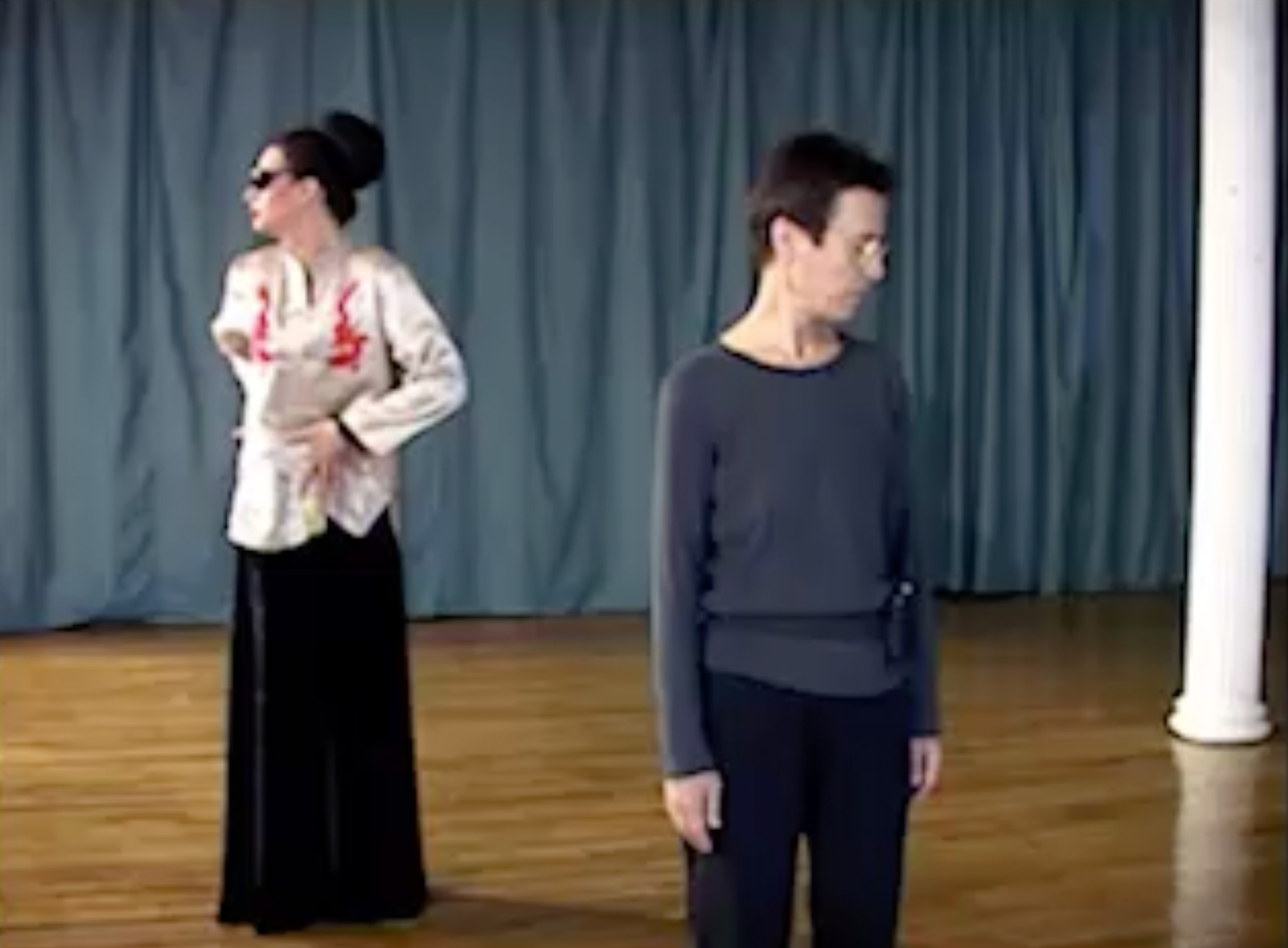 Color video. Rehearsal set in a studio with wooden floors, a blue curtain in the back, and white columns throughout the space. A woman (Yvonne Rainer) wears a long-sleeve shirt and black pants; Charles Atlas wears a floor-length black dress and an embroidered silk top, with hair arranged as a stark, large bun, and dramatic facial make-up. Late in the first part, Atlas wears a shiny, purple dress with a black mid-section and red and gold accents. Most of the second part features a close view of Rainer and Atlas, now in a black bra, laying on the ground, head-to-head, having a conversation.