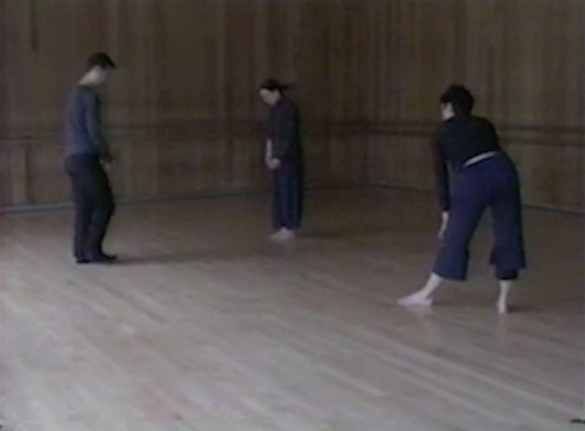 Color video of Rainer observing and instructing two female dancers in a studio space. The barefooted dancers wear dark-colored turtlenecks and loose blue pants. Rainer wears a loose long-sleeve shirt and black pants with dark-colored socks.