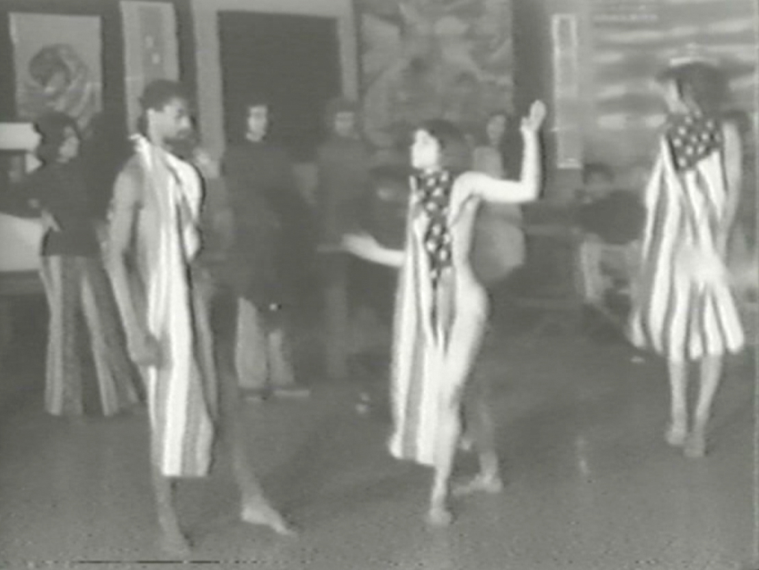 Black-and-white video of the interior of a large hall crowded with people, and with artworks adorning the walls that variously satirize or manipulate the U.S. flag. People mingle amongst themselves until video cuts to performance of Yvonne Rainer’s dance *Trio A with Flags*. Then, individuals including a man wearing a leather jacket (gallerist Stephen Radich), deliver short remarks into a standing microphone.