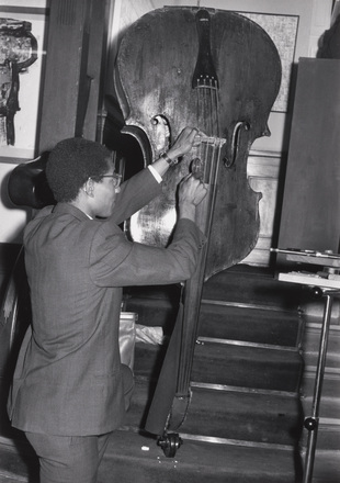 Black-and-white full-body photograph of man in suit turned away from camera to play upright base while holding a hammer. In the background, a man behind lectern on raised stage smiles to camera.