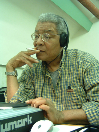 Color photograph of a man sitting man who smokes a cigarette and listens to audio on headphones connected to a Numark audio device. 