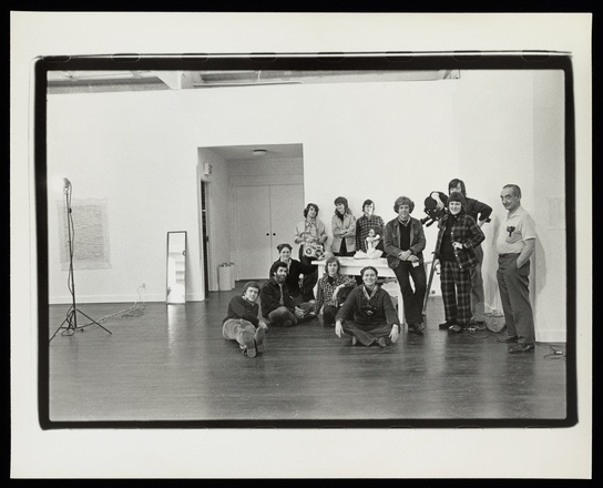 Black and white photograph of a group of people standing in the corner of a white-walled, wooden floored gallery. They candid-appearing shot shows them smiling, and some are in motion as if they are still getting ready for their picture to be taken.