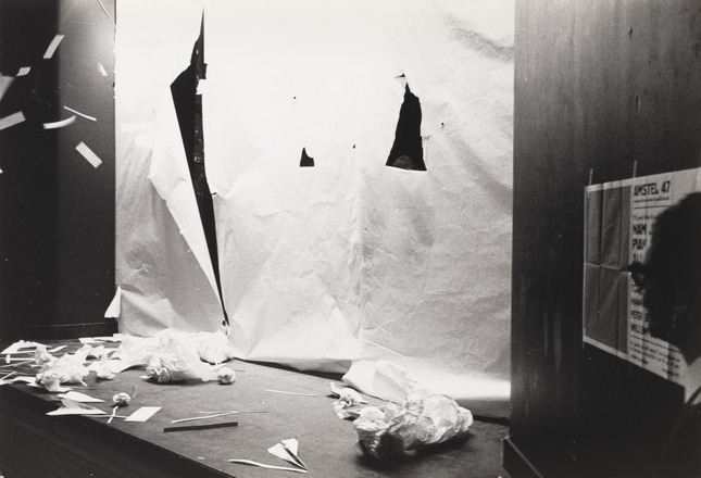 Black-and-white photograph of stage shot from house right. Suspended sheet of white paper spans width of stage and contains one human-sized gash at left and large hole at right. Stage floor in front of sheet strewn with ripped and crumpled paper.