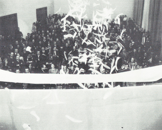 Black-and-white photograph with overhead view of four men punching holes in suspended sheet of paper. At the top of frame, just in view, seated audience faces the opposite side of the sheet.