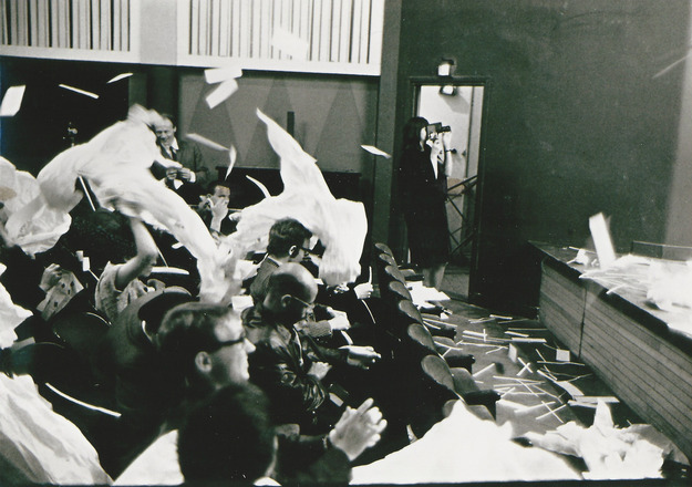 Black-and-white landscape-oriented photograph of people sat in one row of auditorium chairs ripping and tossing paper into the air. Photograph shot from an aisle showing audience from the side. Paper strewn about floor in between chairs and a stage that is just visible at right edge of frame.