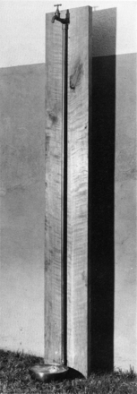 Black and white photograph of outdoor faucet set up in front of a wall. A small metal basin rests on the grass under a spigot mounted high above, attached to the top of a remarkably tall length of pipe that is mounted to an equally tall slab of wood.