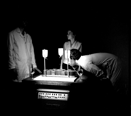 Black-and-white photograph of three people in lab coats surrounding an instrument comprised of three water bottles suspended above three plastic pots that sit on a rectangular box. An amplifier rests below the pots inside the box. The man to the right of the box leans in, bent at the waist, to examine the instrument. The other two figures at center back and left remain upright. The whole scene is lit by a spotlight. 