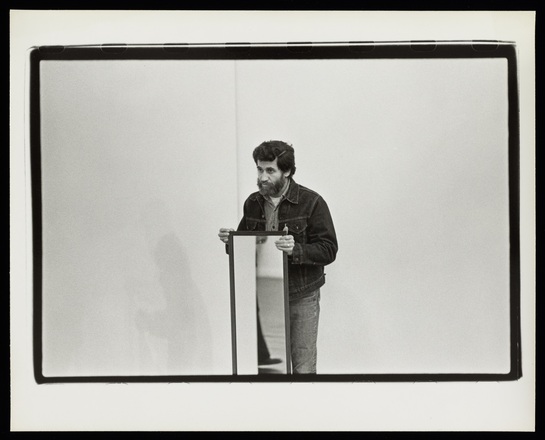Photograph of a man with a beard and wearing a jean jacket standing in the corner of a white room holding the top edges of a full-length mirror which appears to be resting on the floor, which is not visible.