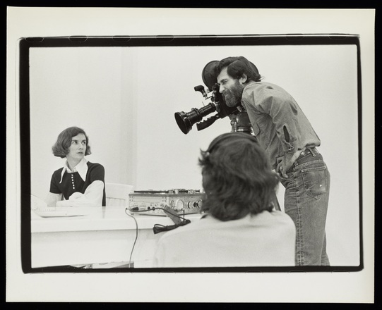 Landscape-oriented black and white photograph of woman sat behind table at left and man with film camera turned to film woman at left. Audio equipment at center on table. Back of head wearing headphones out of focus in central foreground. 