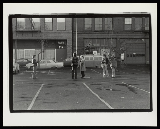 Black and white photograph of a group of five people standing in a parking lot in front of a building designated “PCVA 117.” Two of the figures stand a distance away, holding moving-image recording equipment, including a boom microphone on a long pole.