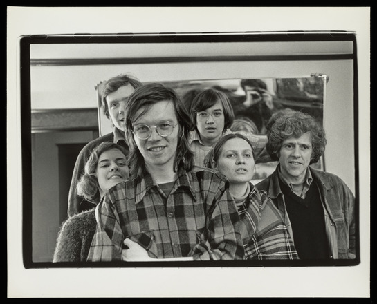 Black and white photograph of a group of six, tightly grouped figures looking into the camera. The photographer is blurrily visible in a mirror held up behind the group by an individual whose hands can only be seen.