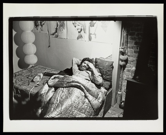 Black and white photograph of a bearded man reclining on a platform bed next to a partially exposed brick wall with a stove in the darkened right corner of the image. Above him on the wall hang a series of four nude or partially nude figure drawings.
