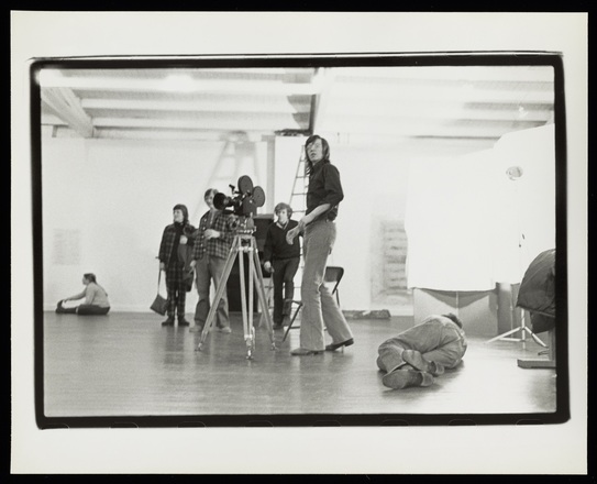 Black and white photograph of six people in a white-walled, wooden floored interior at various distances between far background and close foreground. There is a moving image camera at center, pointed toward the left, out of the frame. Most of the people look in that direction.