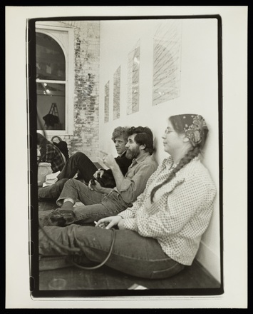 Black and white photograph of three people seated on the floor, leaning against the wall, and two other figures sitting on the floor in the background. There is a far brick wall and darkened window visible, suggesting that it is night.