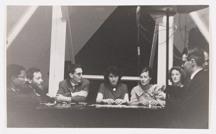 Black-and-white landscape-oriented photograph of four men and three women sat indoors in an arc around a table. Most are looking to the man sat furthest right whose hands are in motion.