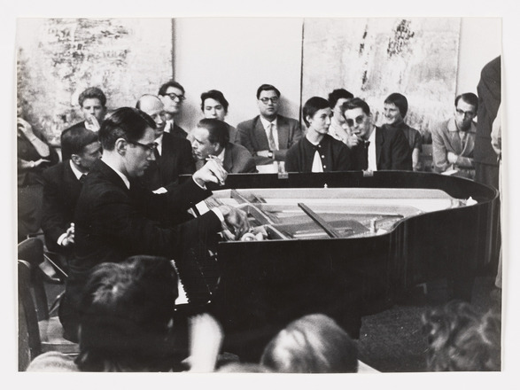 Landscape-oriented black-and-white photograph of a man at left seated behind a grand piano, at right, with its lid removed. The man’s hands are in motion plucking the piano’s strings. Man and piano are surrounded by onlookers in the foreground and background