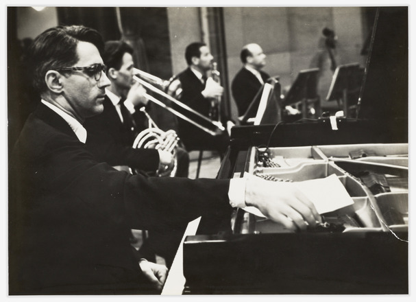 Landscape-oriented black-and-white waist-up photograph of a man at left seated at a piano just visible on the right, with an orchestra in the background. 
