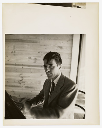 Black-and-white portrait of Tudor in a suit looking directly to the camera while seated at and playing a piano. Soft light from a window at his left touches his back and the left side of his face. Background is dominated by a wall paneled with light-colored wood.