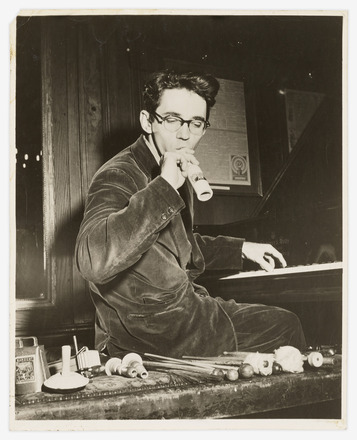 Black-and-white photograph of Tudor sitting at a piano. The man is turned facing the camera, and in front of him is an array of auxiliary percussion instruments arranged on a low bench. His right hand holds an instrument to his mouth, while his left hand reaches back to play the piano.