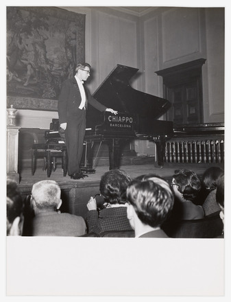 Black-and-white photograph of Tudor in a tuxedo standing on a stage and resting one hand on a grand piano. At the bottom of the frame, the backs of audience members’ heads are visible as they look up toward the stage.