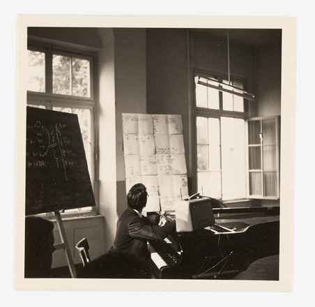 :Square black-and-white photograph of a man, at center, seated at a grand piano and facing away from the camera and looking at a tall rectangular board filled with eighteen papers arranged in a grid. Each page contains different handwritten markings in black. A chalkboard with notes is to the man’s left. An open, light-filled window is at the right of the frame. 