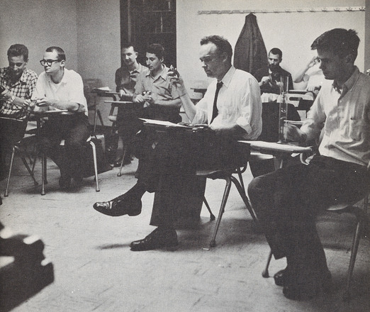 Black and white photograph of eight men in a classroom, each sat at a chair desk. Photograph shot at eye level of sitting figures.