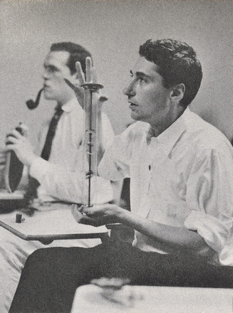 Portrait-oriented black and white photograph of one man shot from the left sitting at a desk while holding a plastic tube. Another man behind sitting, smoking a pipe, and holding a toy ship. 