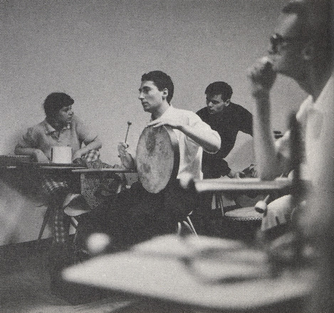 Square black and white photograph of a man shot from the front left sitting at a desk while hitting a handheld drum. Two men behind sitting at desks. One blurry man and desk in foreground framing the scene.