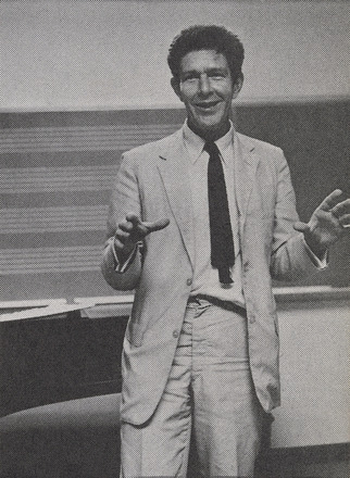 Portrait-oriented black and white photograph of a man shot from knees up in a tan suit and black tie smiling and gesturing. Chalkboards on wall behind.