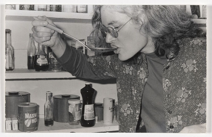 Landscape-oriented black and white photograph of a woman at right shot from chest up eating from a long serving spoon. Stocked pantry shelves in background at left. 