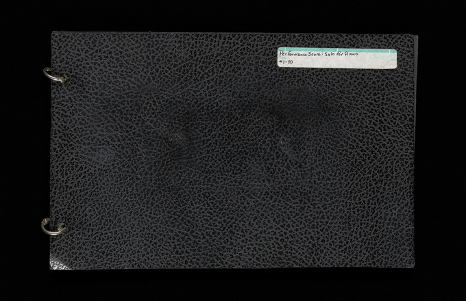 Horizontal rectangular two-ring binder with black cover containing forty cream pages with two hole punches at leftmost edge. White sticker with black handwritten text reading “Performance Score: Solo for Piano # 1-80” at top right of front cover. Pages are photographed in two-page spreads, with hole-punched ends connected at center to binder rings. Pages contain graphic notation in black pen and pencil. Each page contains four vertical line segments roughly the height of a staff, two each at left and right edge,s to create two horizontal bands of space across the page. Notations on pages in line with bands are either (1) dots attached to rectangular boxes with text by a vertical line, (2) a horizontal staff segment with at minimum one clef and one musical note, or (3) Y-shaped markings annotated with letters. Most pages contain five or fewer groupings of notations. Many notations are annotated with either text or numbers. Rightmost edges of pages are annotated in pencil with numbers. Paginated 1–90 at bottom right of each page. Final two pages contain a handwritten list of materials.