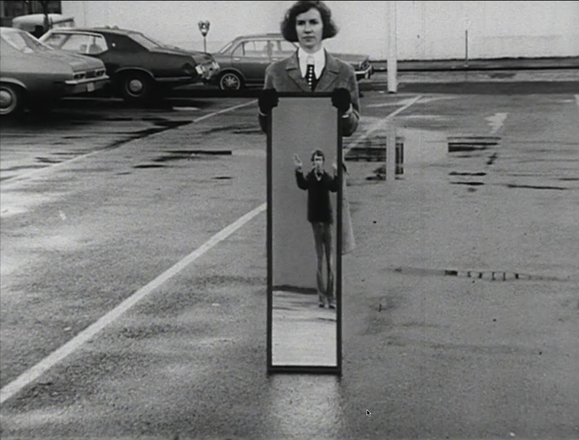 Landscape-oriented black and white photograph with a woman at the center of the frame holding a full-body mirror outdoors in a parking lot. Reflection shows man with hands raised overhead. 