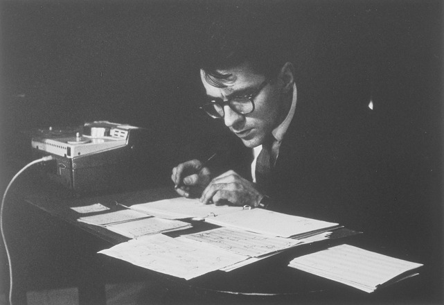 Landscape-oriented black-and-white photograph of a man, at center, with his face visible to the camera while he sits behind a table covered with staff sheets. The man’s hands are on table, one holding a pen, the other flipping through the sheets.