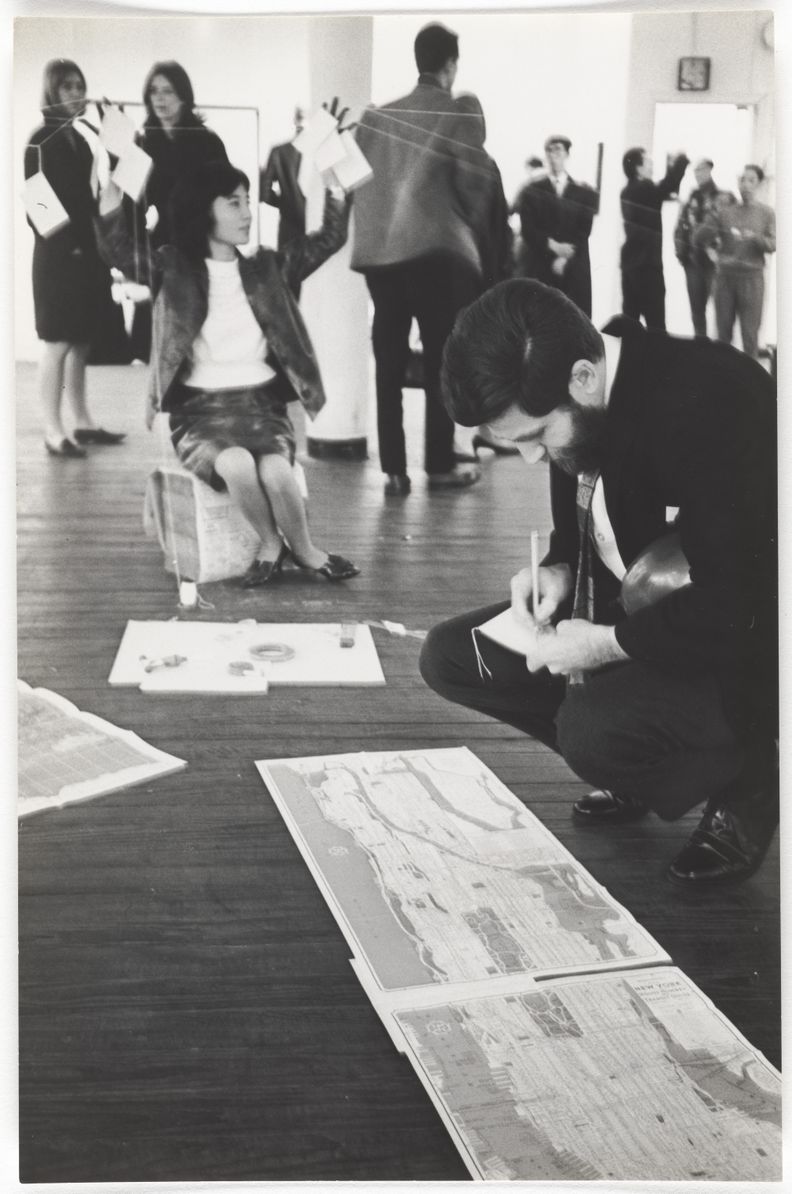Black-and-white photograph of a woman sitting on a box in the middle of a room raising her left hand from which thin wires suspend out of frame. The woman is surrounded by people in the background, while in the foreground a suited man kneels, writing and looking at a map on the floor.