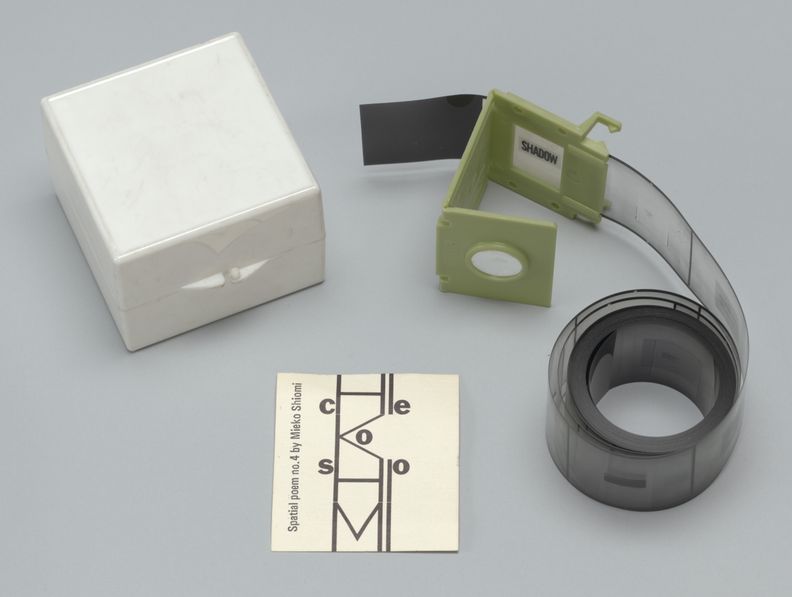 Three objects against a gray background. One is a white box with a hinge. To the right is a roll of microfilm placed in a green plastic viewing device. At the bottom is a small piece of paper with text, Spatial poem number 4 by Mieko Shiomi, above a pattern adorned with the letters C, S, E, O and O.