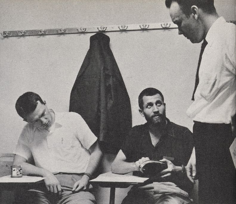 Landscape-oriented black and white photograph of three men, two sitting at desks on left, and one standing at right, all conversing indoors.