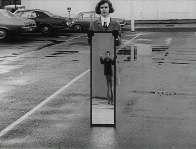 Landscape-oriented black and white photograph with a woman at the center of the frame standing in a wet parking lot holding a full-body mirror outdoors in a parking lot. Reflection shows a man with hands raised overhead.