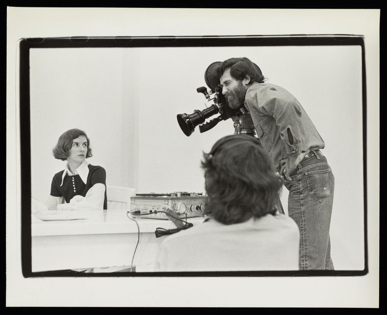 Landscape-oriented black and white photograph of a woman sitting behind a table at left while a man films her with a camera. There is audio equipment at center on table, and the back of the head of a man wearing headphones is out of focus in the central foreground.