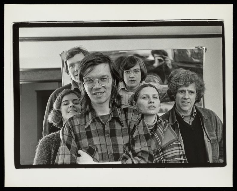 A landscape-oriented black and white photograph of six people clustered together and smiling to the camera. They are shot from the chest up.