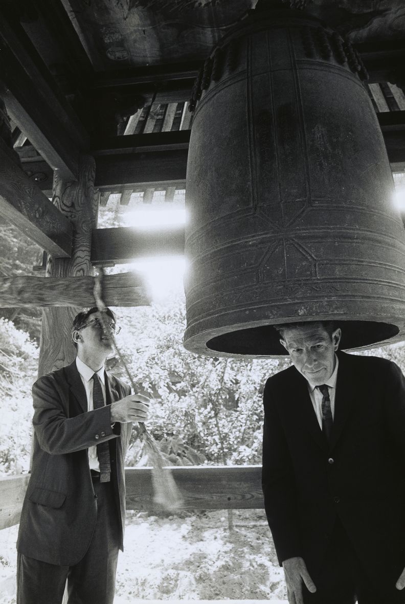 Portrait-oriented black-and-white photograph of two men under a pavilion. The man at right stands with his head inside a large bell, and the man at left moves to hit the bell with a mounted wood hammer.