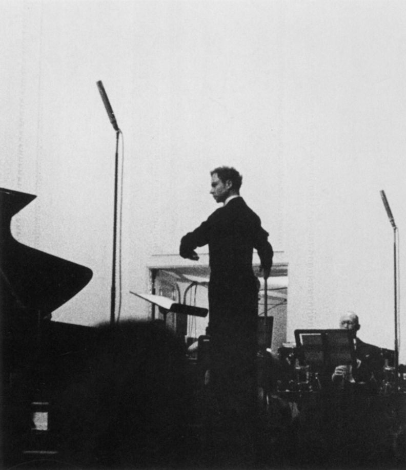 Square-shaped black-and-white photograph of a man on a stage turned away from the camera and conducting an orchestra. The photo was shot from the audience.