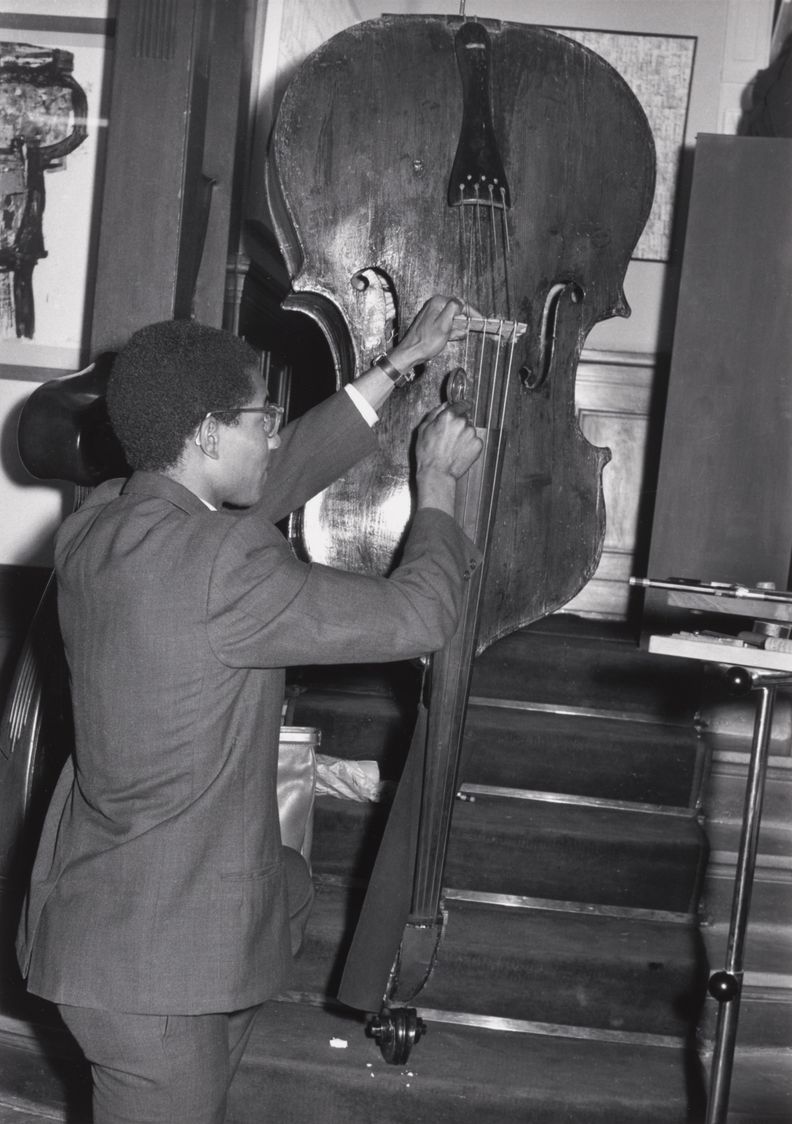 Black-and-white full-body photograph of man in suit turned away from camera to play an upside down upright bass using an implement in his right hand. The scroll of the bass sits on the bottom stair of a flight of stairs leading to a wall.
