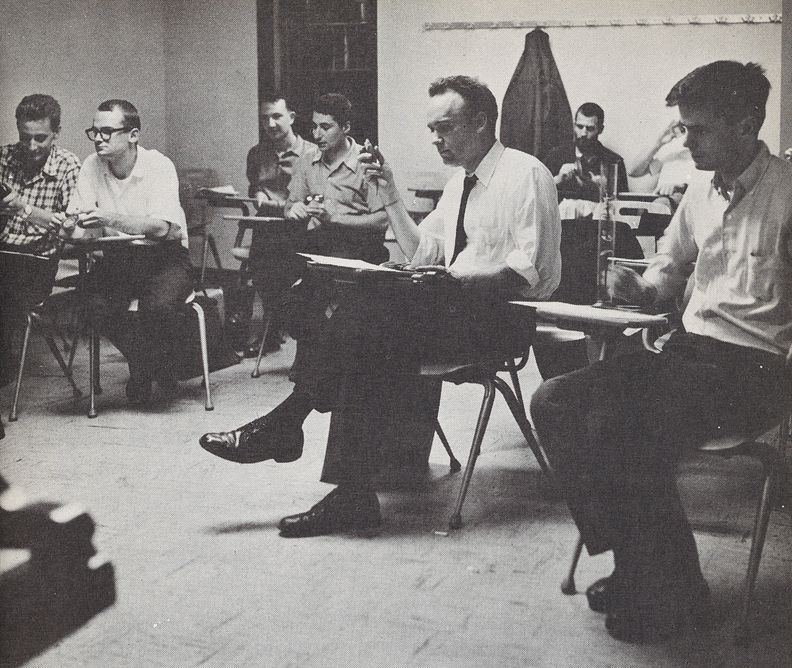 Black-and-white photograph of eight men in a classroom, each sat at a chair desk. Photograph shot at eye level of sitting figures.
